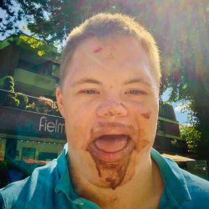 Ein junger Mann ist frontal im Freien fotografiert und blickt mit geöffnetem Mund in die Kamera. Im Mund- und Kinnbereich sind braune Verschmutzungen zu sehen. Im Hintergrund befinden sich Bäume und ein Gebäude mit teilweise sichtbarer Beschriftung.