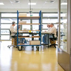 Mehrere Personen sitzen in einem hellen Arbeitsraum an Tischen und verrichten Tätigkeiten an Arbeitsplätzen. Sie tragen weiße Arbeitskleidung, Hauben und sitzen auf Bürostühlen. In der Mitte des Raumes steht ein Regalwagen mit Kunststoffboxen, im Hintergrund sind Fenster und weitere Arbeitsplätze zu sehen.
