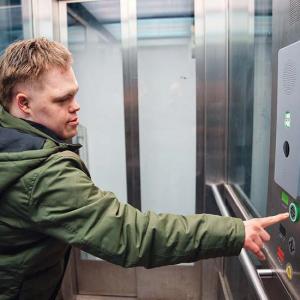 Eine Person in einer grünen Jacke steht in einem Aufzug und drückt mit ausgestrecktem Arm einen Knopf an der Bedienleiste. Die Aufzugskabine hat Metallwände und seitliche Glasflächen. Auf der Leiste sind mehrere Tasten, ein Lautsprecherfeld und kleine Beschriftungen zu sehen.