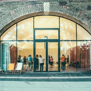 Die Aufnahme zeigt die Fassade eines Backsteingebäudes mit einem großen, bogenförmigen Fenster- und Türelement. Durch die Glasfront sind mehrere Personen in einem hellen Innenraum zu sehen, einige stehen paarweise oder in kleinen Gruppen. Vor dem Gebäude stehen drei Liegestühle, an den Seiten der Fassade sind Graffiti angebracht.