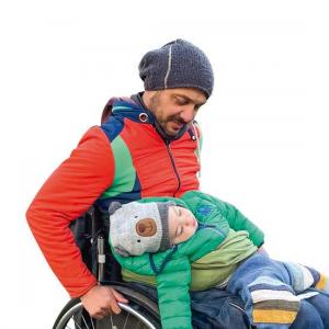 Ein Mann in bunter Jacke und Mütze sitzt in einem Rollstuhl. Auf seinem Schoß liegt ein Kind in grüner Jacke und grauer Mütze mit geschlossenen Augen.