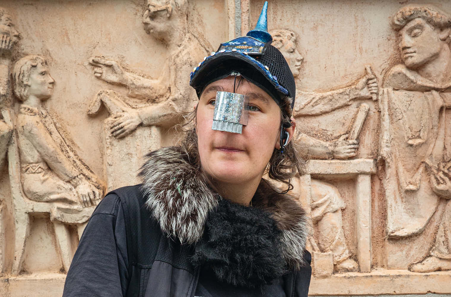 Eine Person trägt eine schwarze Kopfbedeckung mit blauem, spitzem Aufsatz. Auf der Nase befindet sich ein silberfarbenes, rechteckiges Element. Im Hintergrund ist eine Steinwand mit reliefartigen Figuren zu sehen.
