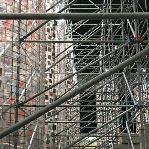 Eine große, begehbare Konstruktion aus zahlreichen grauen Metallrohren füllt den Raum. Dazwischen befinden sich hohe, netzartige Wände mit vielen kleinen rechteckigen Flächen in unterschiedlichen Farben und Materialien; im Hintergrund sind schwarze Treppen und weitere Metallstreben zu sehen.