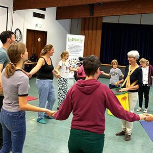 Männer, Frauen und Kinder bewegen sich an Händen haltend im Kreis. Eine Frau in der Mitte des Kreises schlägt eine Trommel.