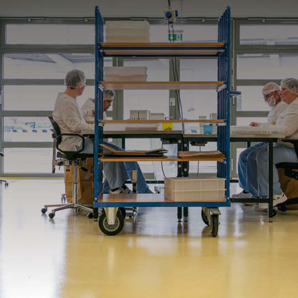 Mehrere Personen sitzen in einem hellen Arbeitsraum an Tischen und verrichten Tätigkeiten an Arbeitsplätzen. Sie tragen weiße Arbeitskleidung, Haub...