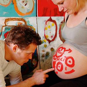 Sanft mit dem Zeigefinger bemalt der Partner von Birte Müller deren hochschwangeren nackten Bauch mit einem lachenden Gesicht, dem Symbol für männl...