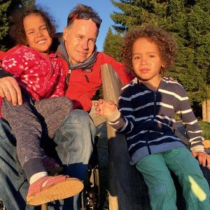 Sebastian Ruppe im Rollstuhl hat seine fünfjährige Tochter Valentina auf seinem Schoß, der Zwillingsbruder Paul sitzt daneben auf einer Bank.