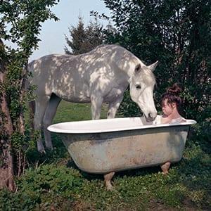 Sofi sitzt im Freien in einer freistehenden Badewanne, ein Pferd steht daneben und trinkt aus der Badewanne.