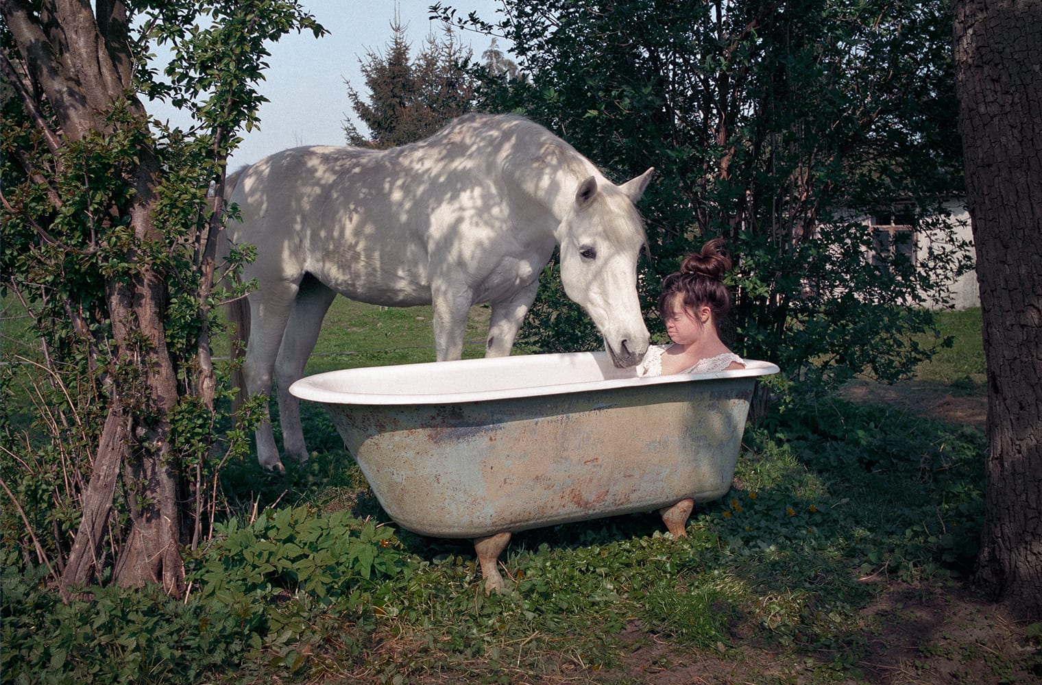 Sofi sitzt im Freien in einer freistehenden Badewanne, ein Pferd steht daneben und trinkt aus der Badewanne.