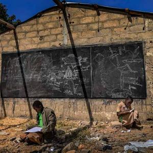 Zwei Kinder sitzen in einem Schulgebäude ohne Dach am Boden und schreiben in ihre Hefte. Im Hintergrund hängt eine mit Kreide beschriebene Schultafel.