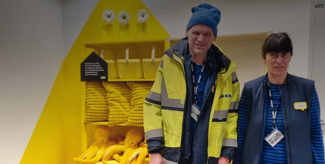 Zwei Mitarbeitende von IKEA stehen vor einer gelben Wand mit Regalen, die mit gelben Stofftieren und Kissen gefüllt sind. Beide tragen Arbeitskleid...