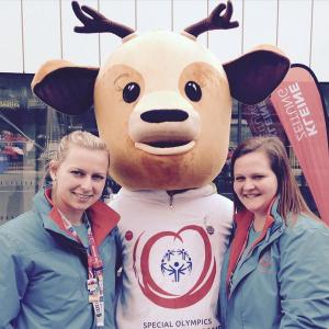 Zwei Studentinnen haben das überlebensgroße Maskottchen der Special-Olympics-Weltwinterspiele 2017 in ihre Mitte genommen und lächeln in die Kamera...